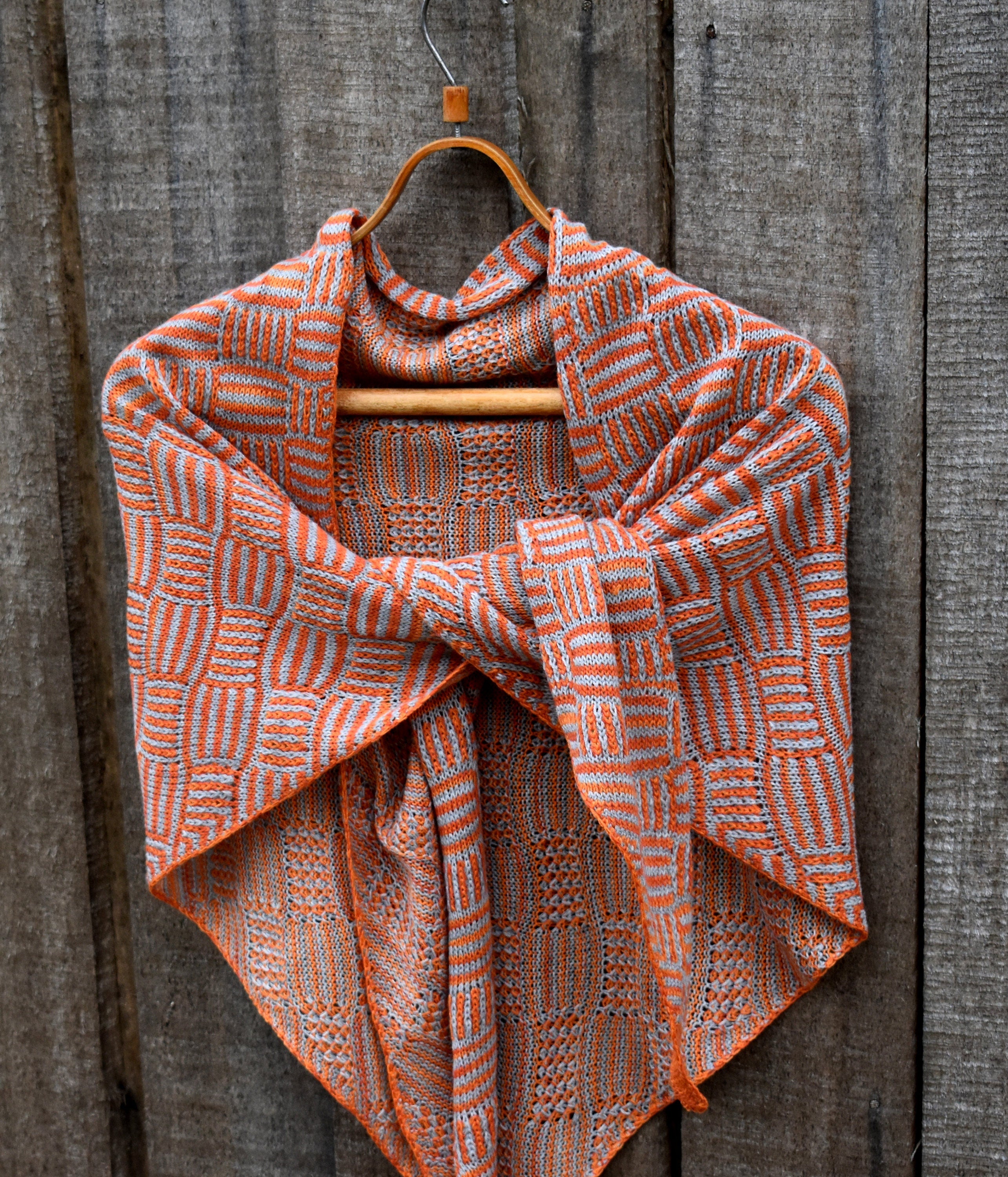 Knitted shawl with orange and white pattern draped over a wooden hanger against a wooden background