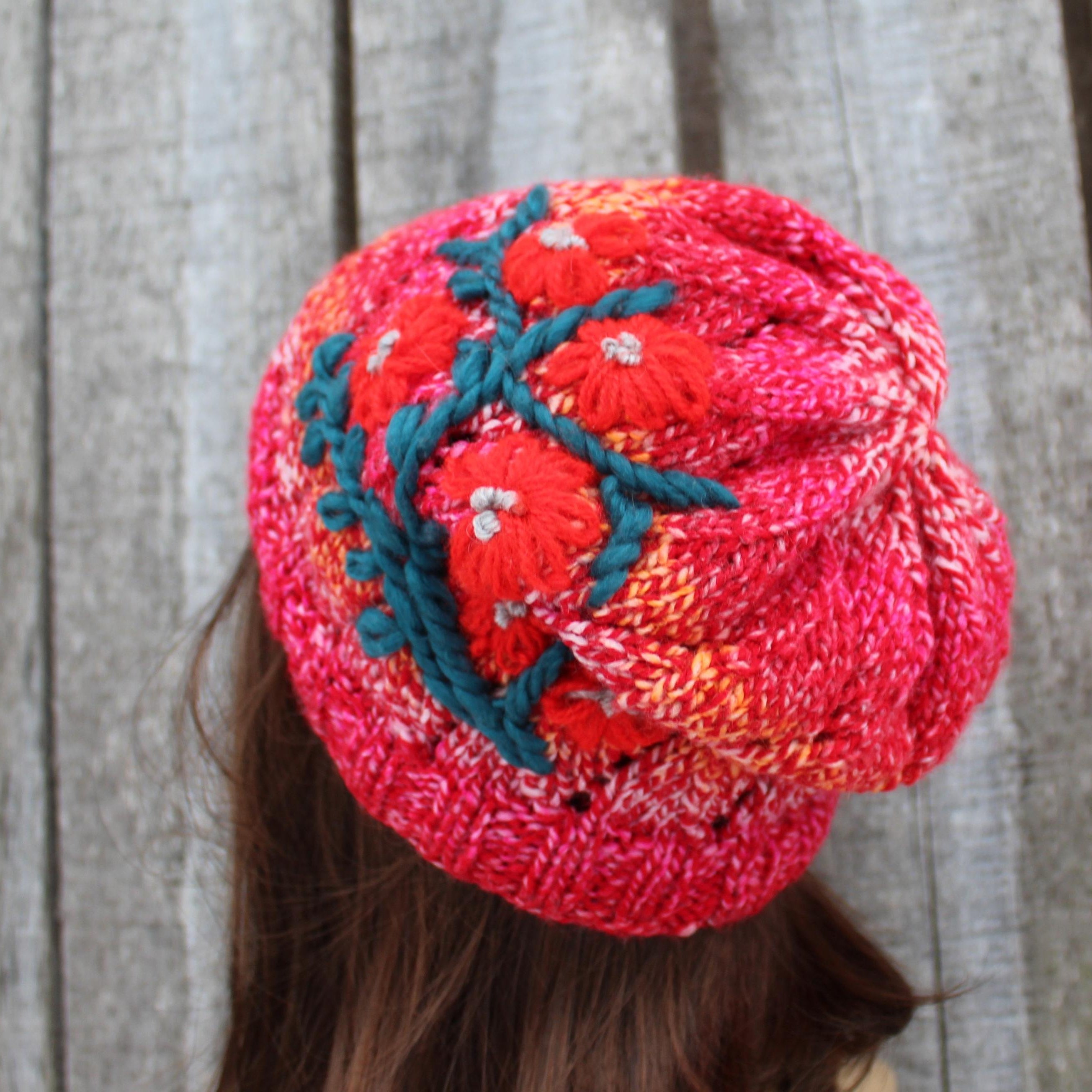 Red knitted beanie with green floral pattern worn by a person against a wooden background