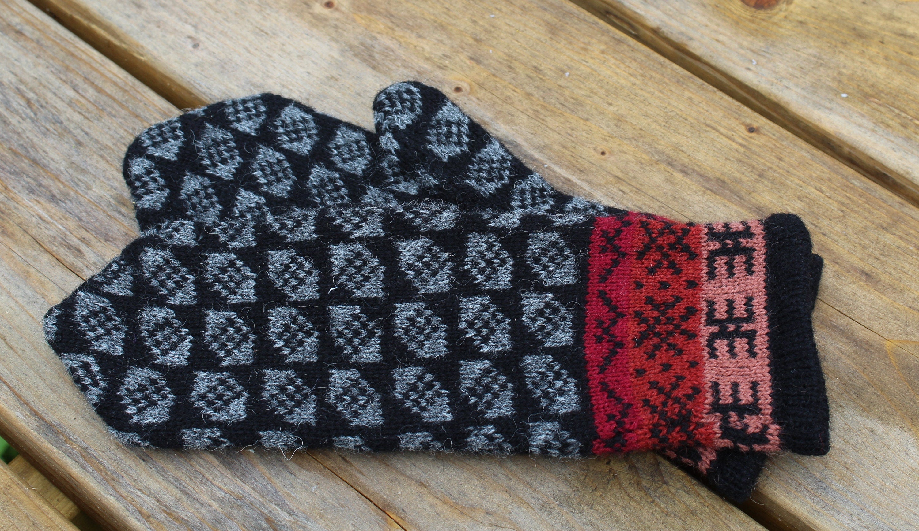 Pair of black and gray patterned mittens with red cuffs on a wooden surface