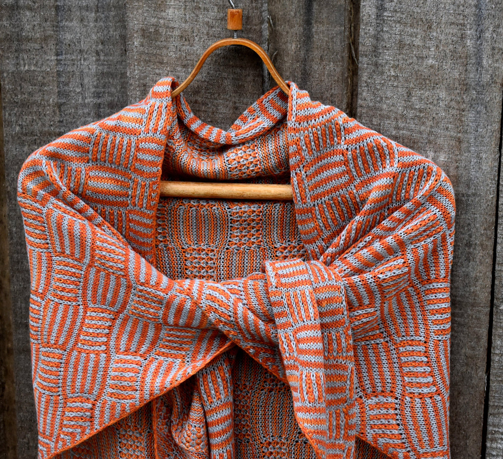Knitted shawl with orange and white pattern draped over a wooden hanger against a rustic wooden background.
