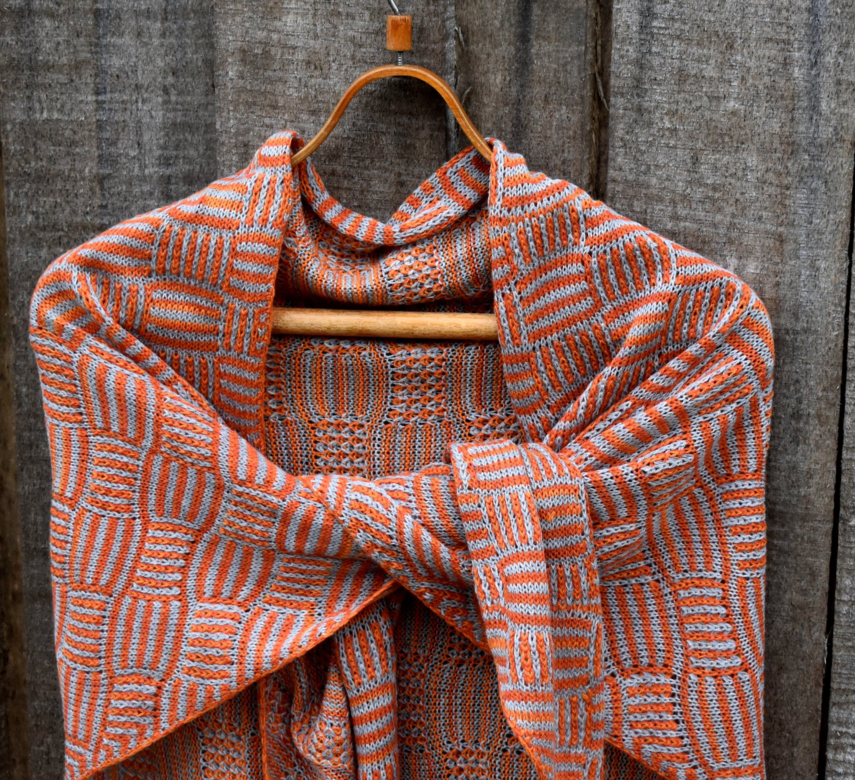 Knitted shawl with orange and white pattern draped over a wooden hanger against a rustic wooden background.