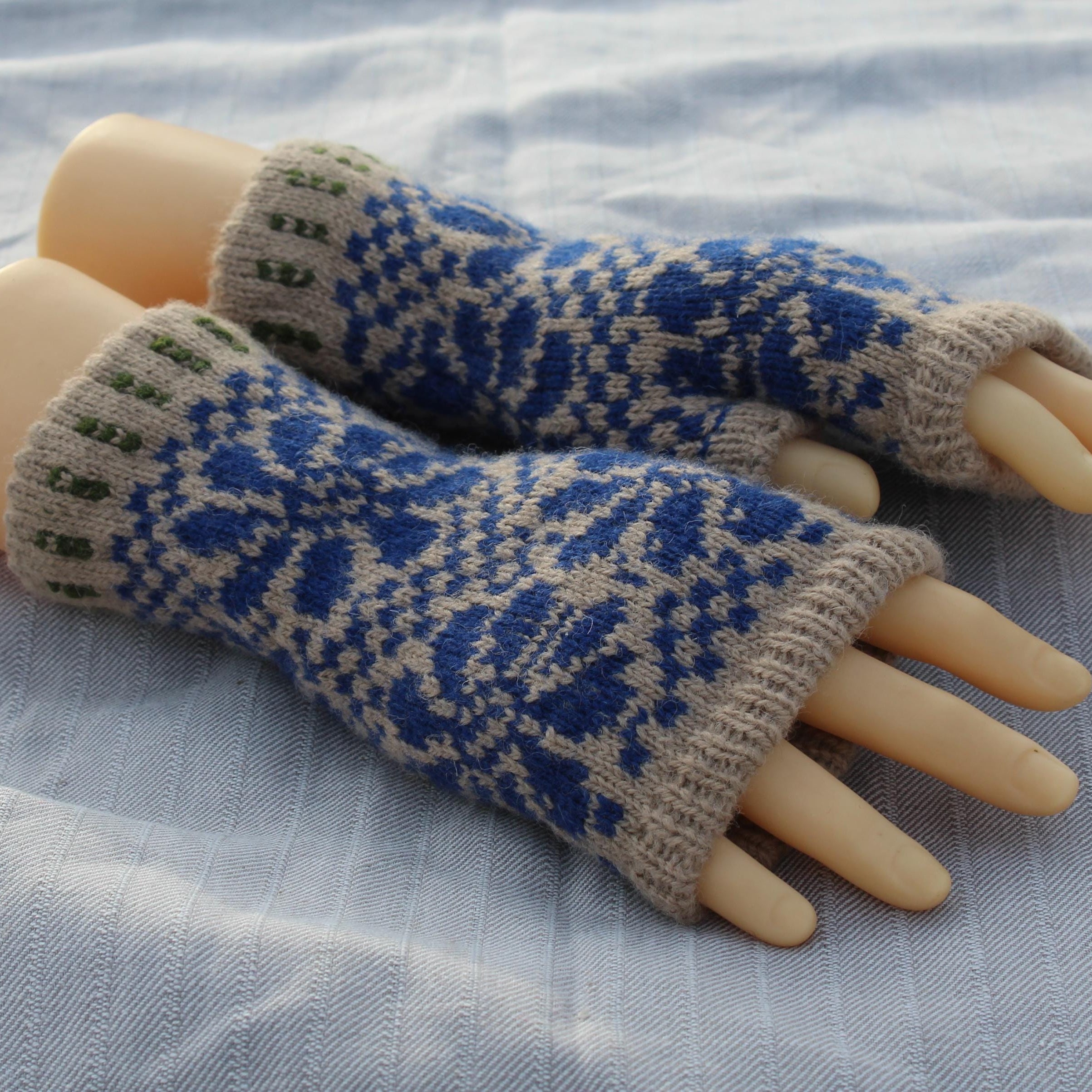 Pair of blue and beige knitted fingerless gloves on a mannequin hand against a light fabric background.