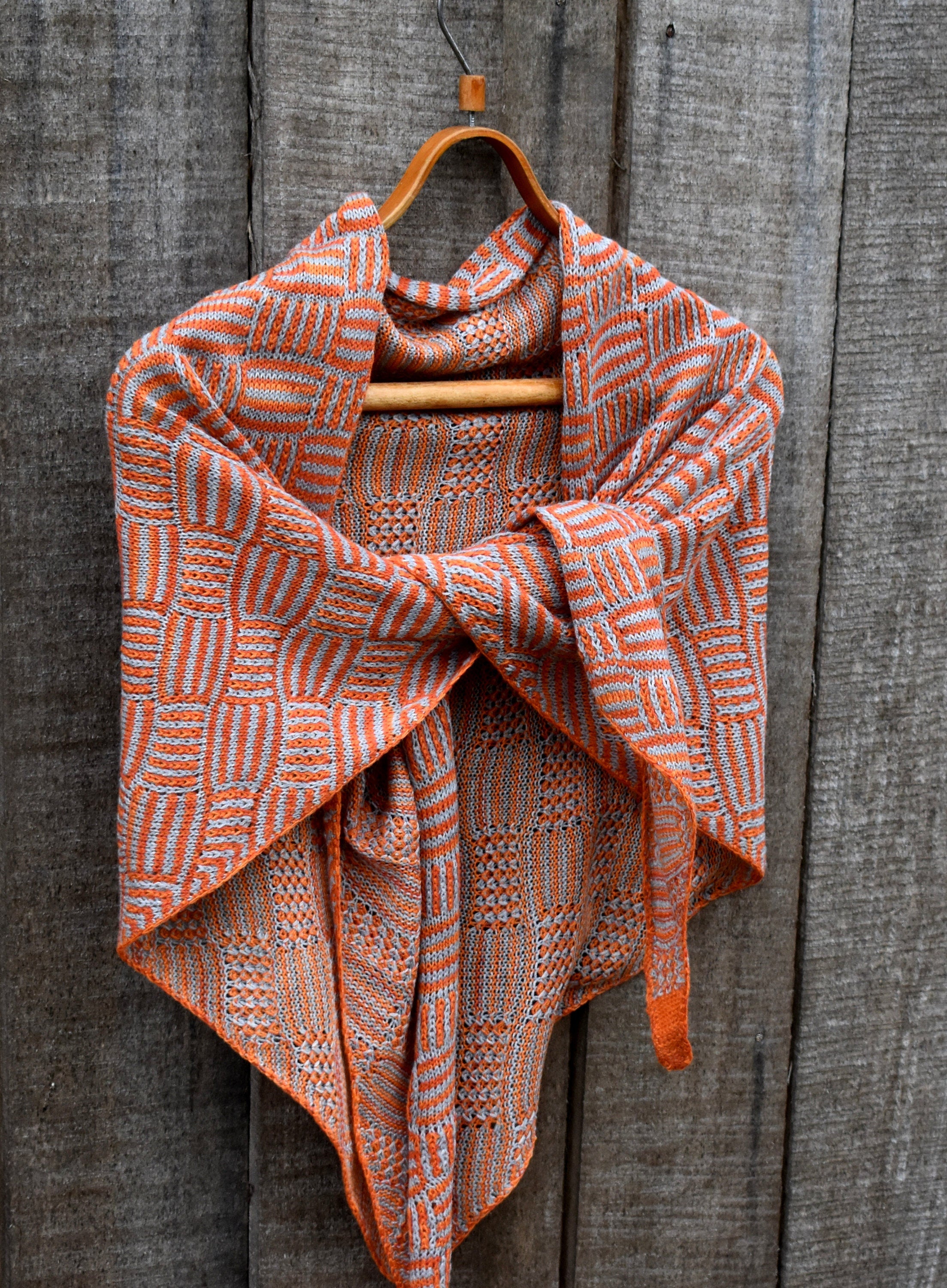 Knitted shawl with orange and white pattern draped over a wooden hanger against a wooden background