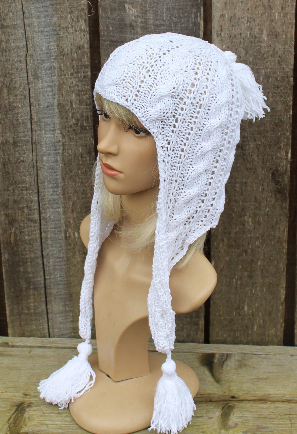 White knitted hat with ear flaps and tassels on a mannequin head against a wooden background