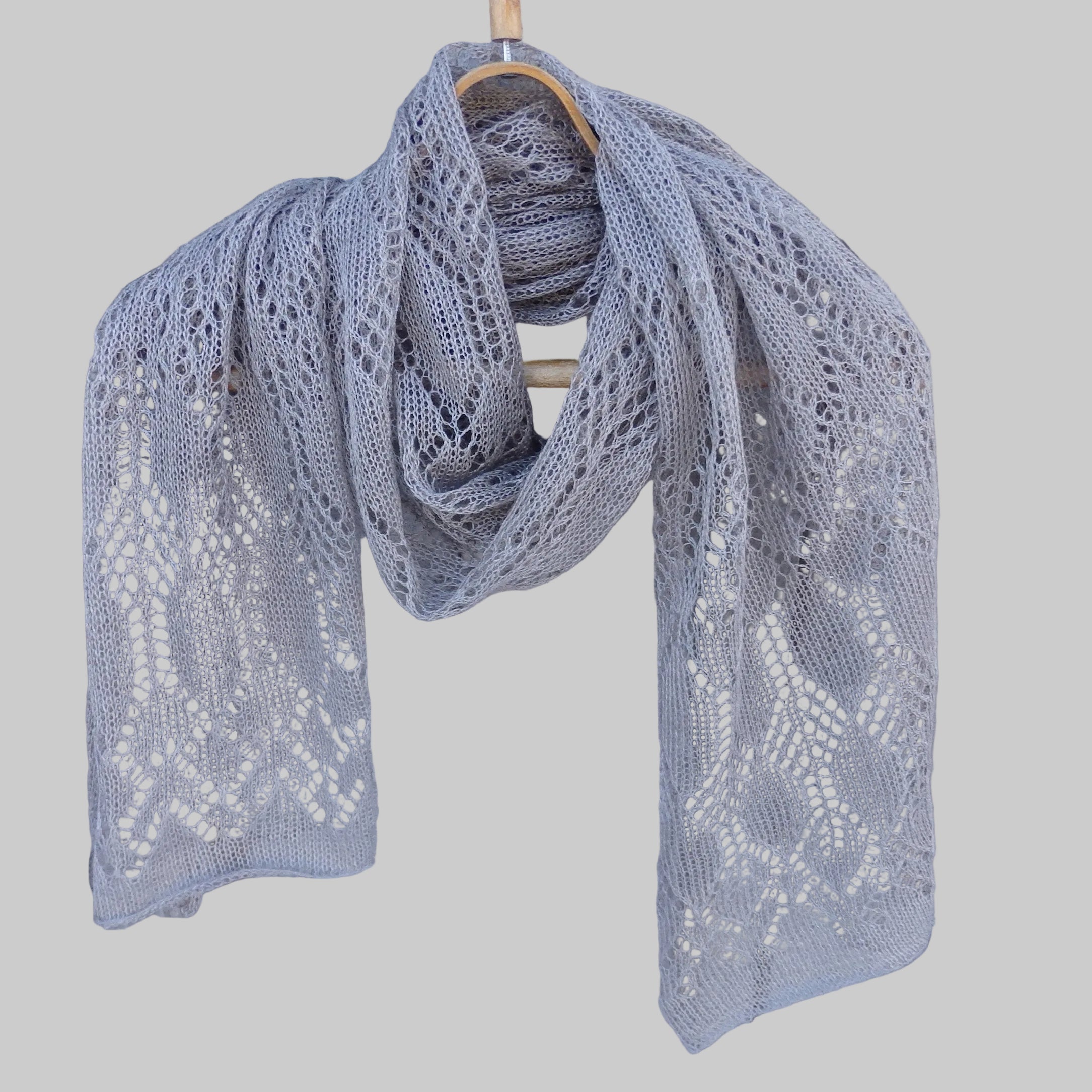 Gray knitted shawl with lace pattern draped over a wooden hanger against a wooden background