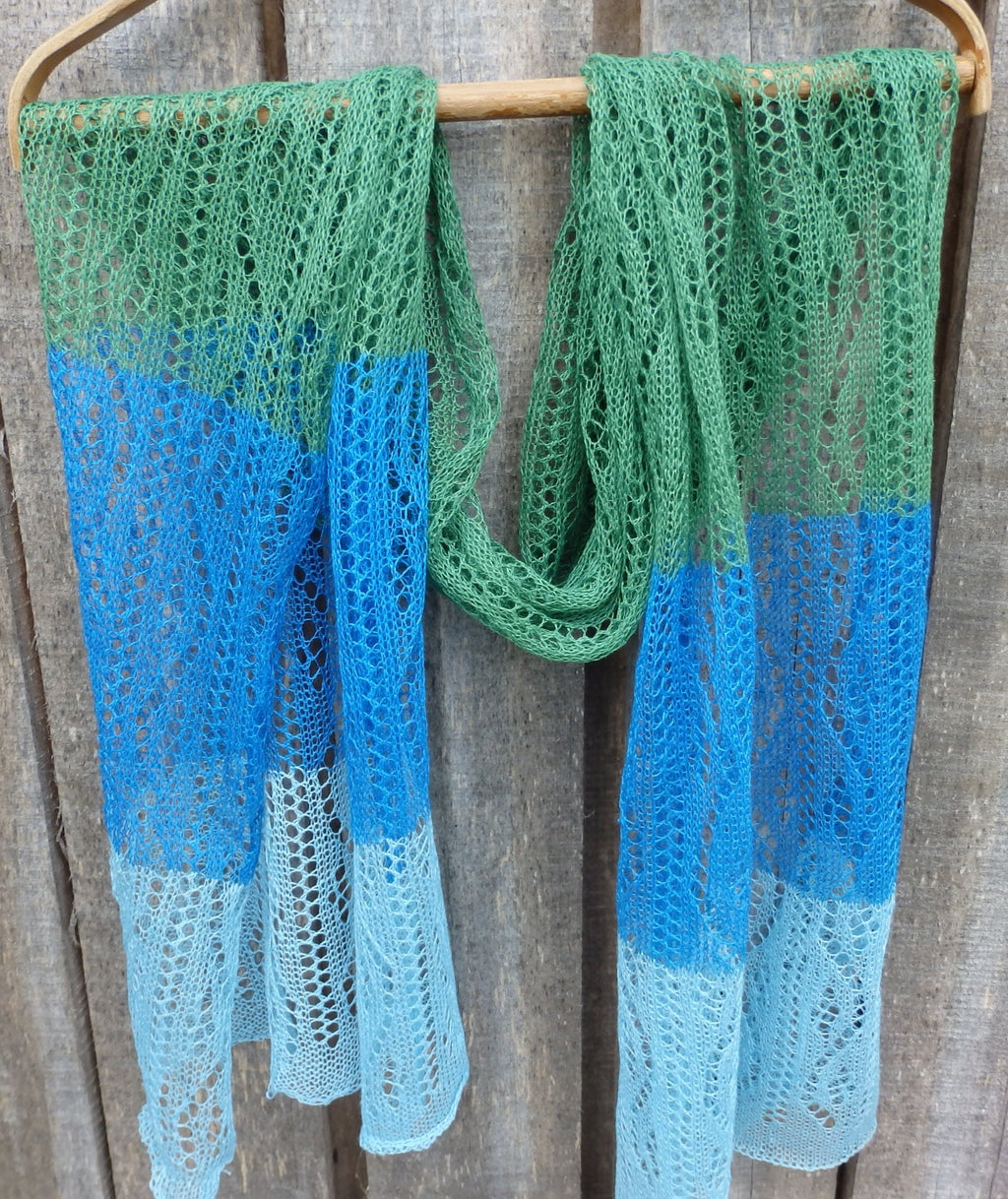 Knitted scarf with green, blue, and light blue gradient draped over a wooden rod against a wooden background.