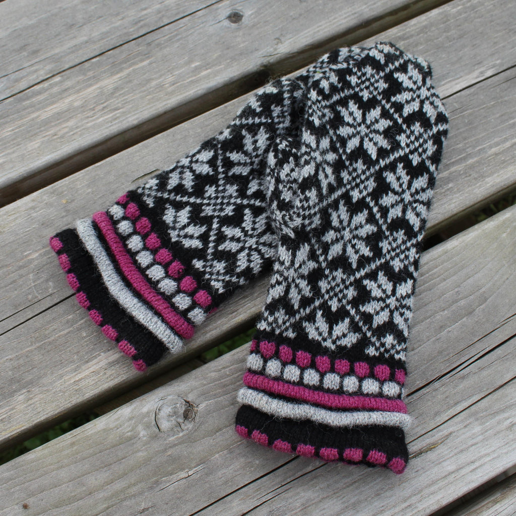 Pair of knitted mittens with a black, white, and pink pattern on a wooden surface.