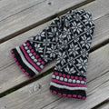 Pair of knitted mittens with a black, white, and pink pattern on a wooden surface.