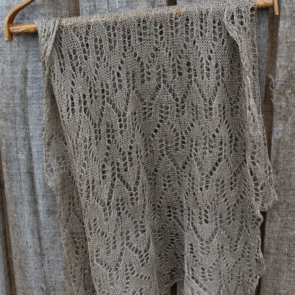 Knitted shawl with a lace pattern draped over a wooden hanger against a rustic wooden background.