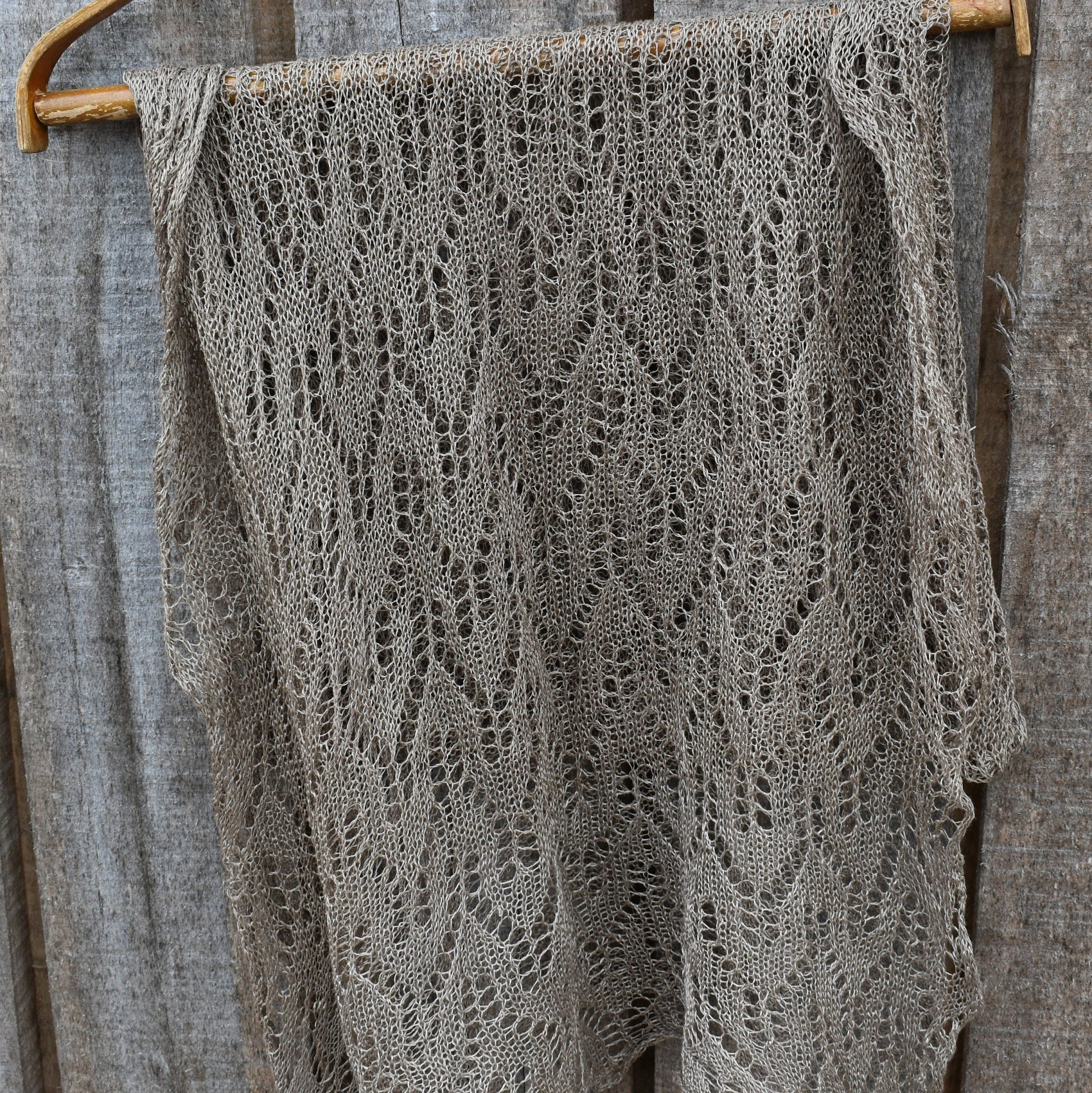 Knitted shawl with a lace pattern draped over a wooden hanger against a rustic wooden background.