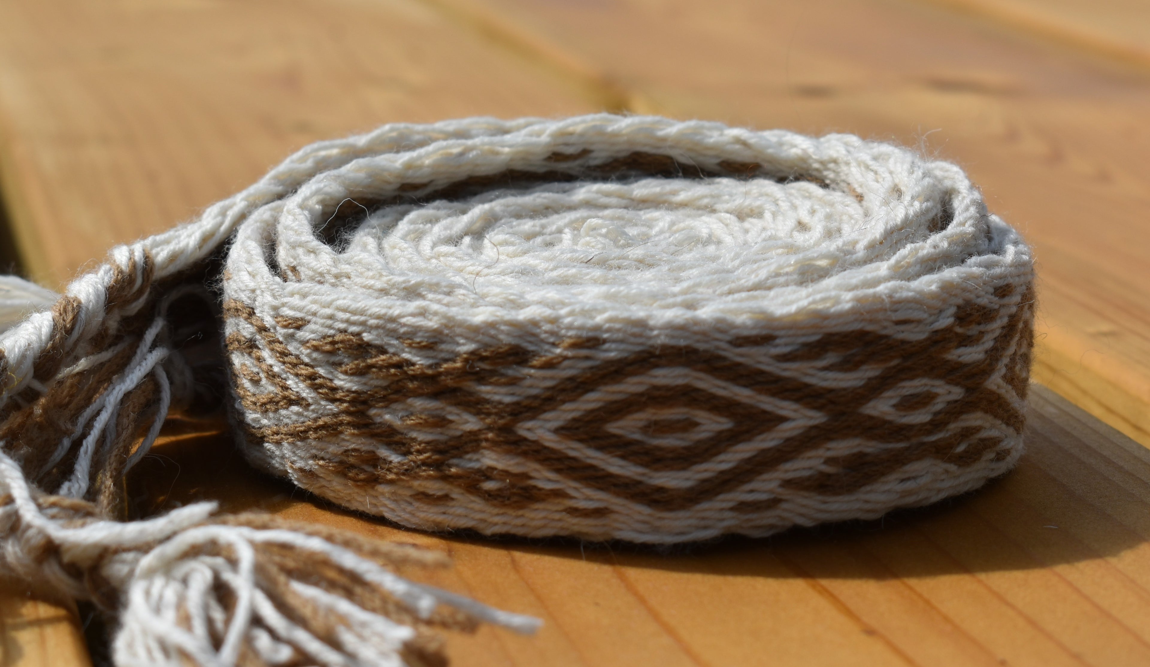 A rolled woven textile with tassels, featuring a geometric diamond pattern in brown and off-white, resting on a wooden surface.