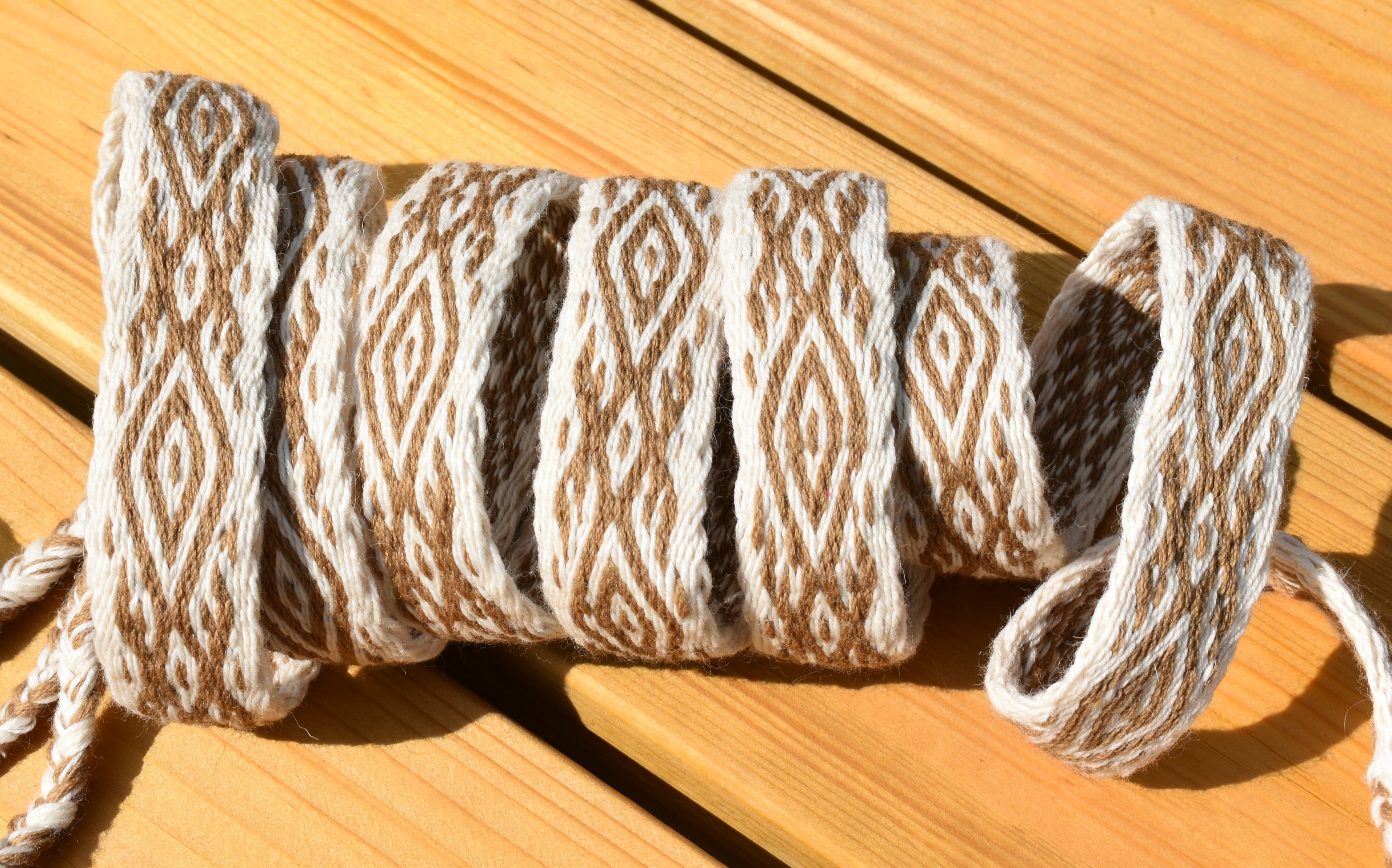 A coiled bundle of braided rope or cord with a repeating diamond pattern in beige and off-white, resting on a wooden surface.