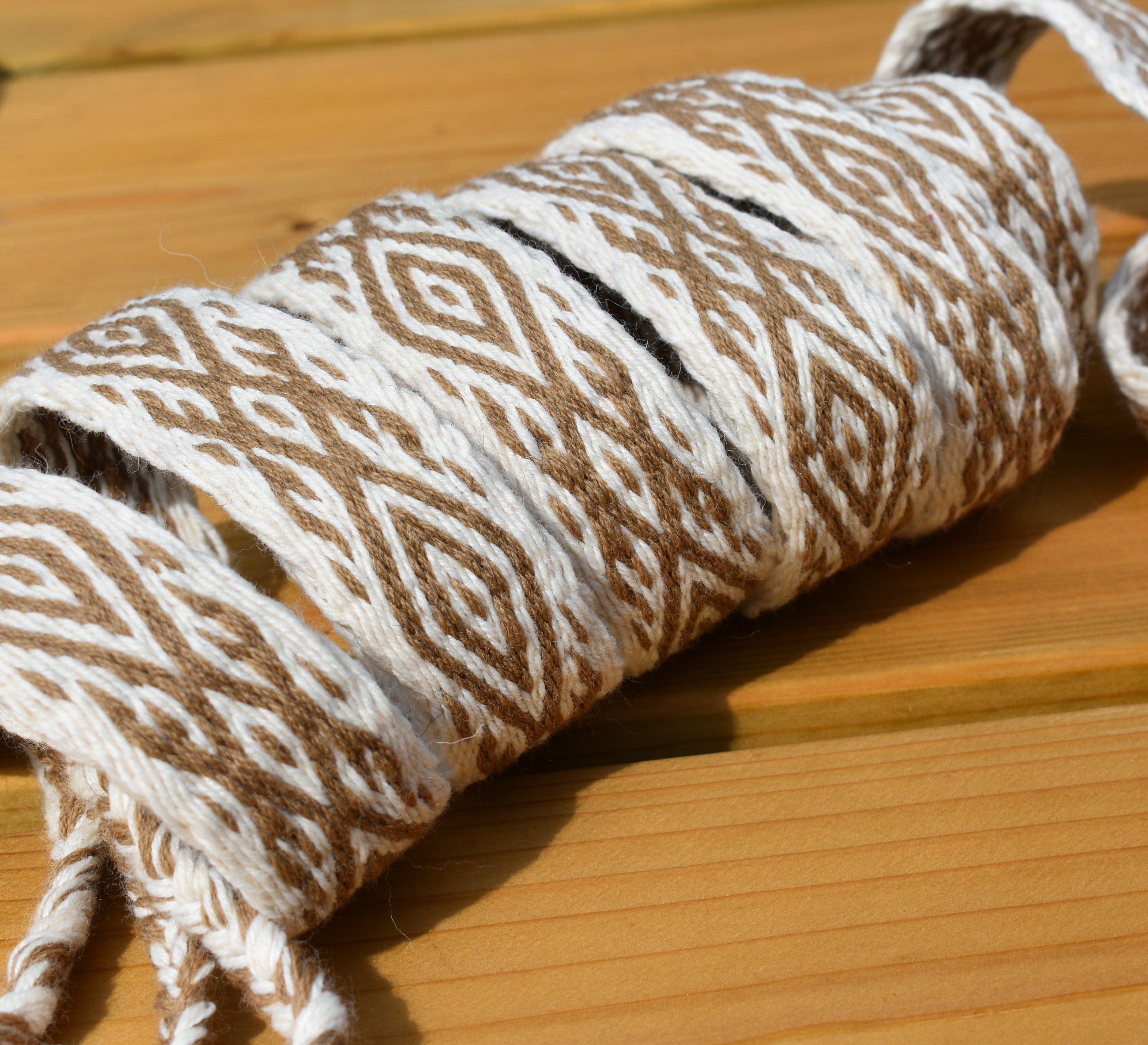 A close-up of a rolled piece of braided twine or cord with a geometric diamond pattern in brown and off-white, resting on a light wood surface.