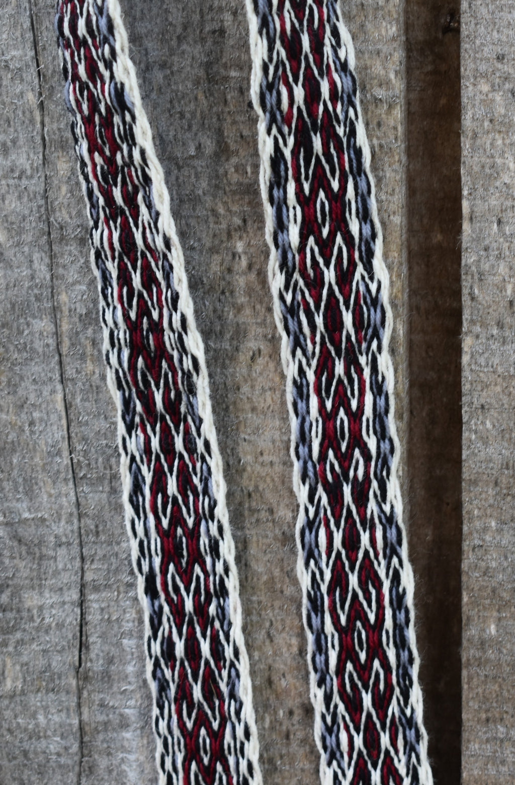 A close-up of a woven textile with two vertical straps or panels featuring an intricate geometric pattern in deep red, off-white, and dark gray on a textured gray fabric background.