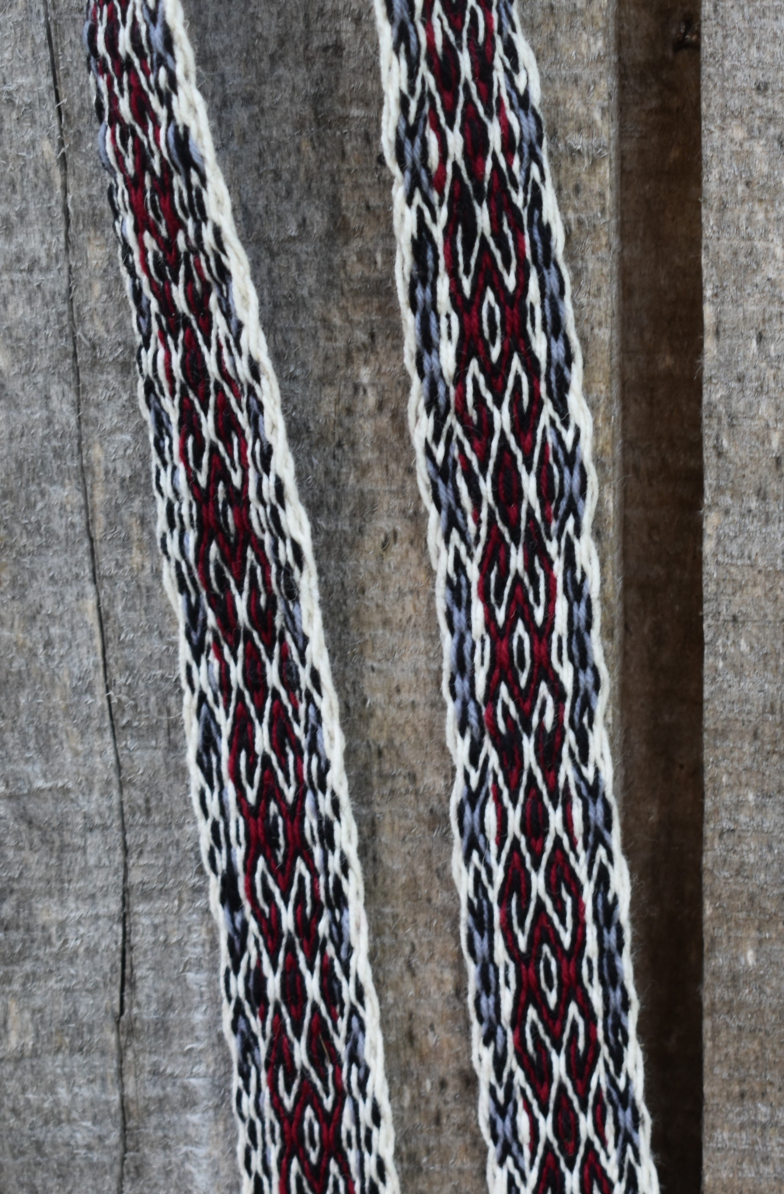 A close-up of a woven textile with two vertical straps or panels featuring an intricate geometric pattern in deep red, off-white, and dark gray on a textured gray fabric background.
