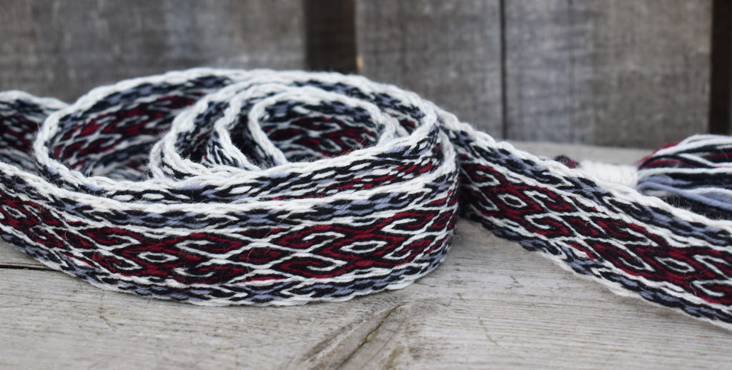 A close-up of a woven strap or belt with a geometric pattern in white, black, and dark red, resting on a rustic wooden surface.