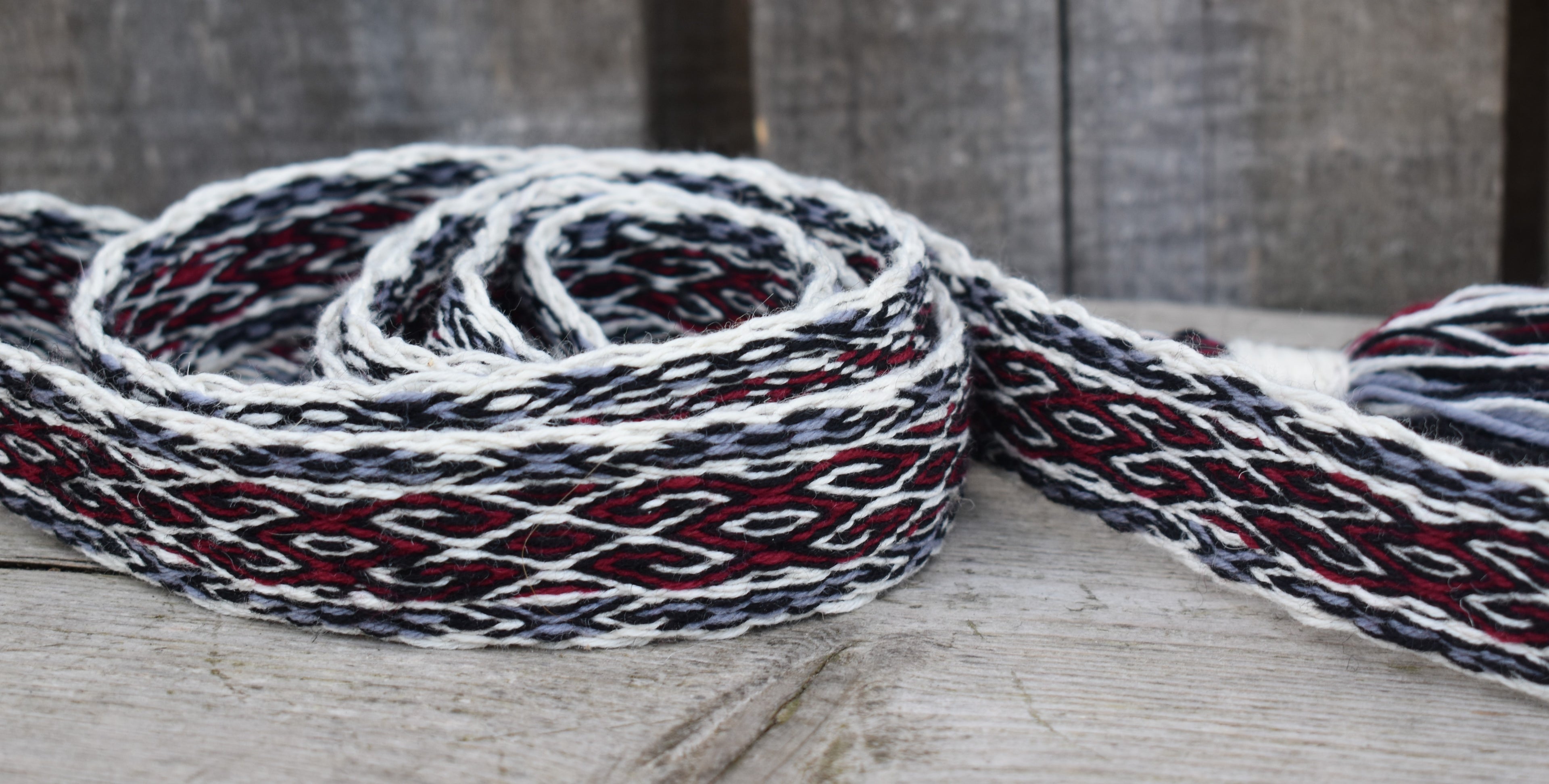 A close-up of a woven strap or belt with a geometric pattern in white, black, and dark red, resting on a rustic wooden surface.