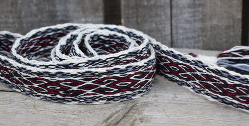 A close-up of a woven strap or belt with a geometric pattern in white, black, and dark red, resting on a rustic wooden surface.