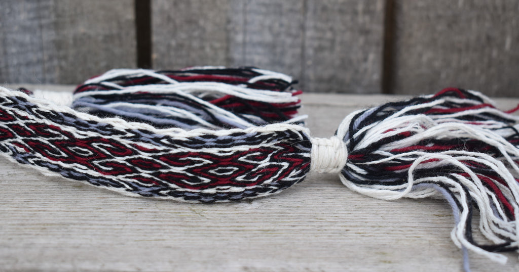A close-up of a braided cord or strap with tassels, made from white, black, and red yarns, resting on a light wooden surface.