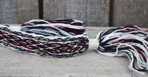A close-up of a braided cord or strap with tassels, made from white, black, and red yarns, resting on a light wooden surface.