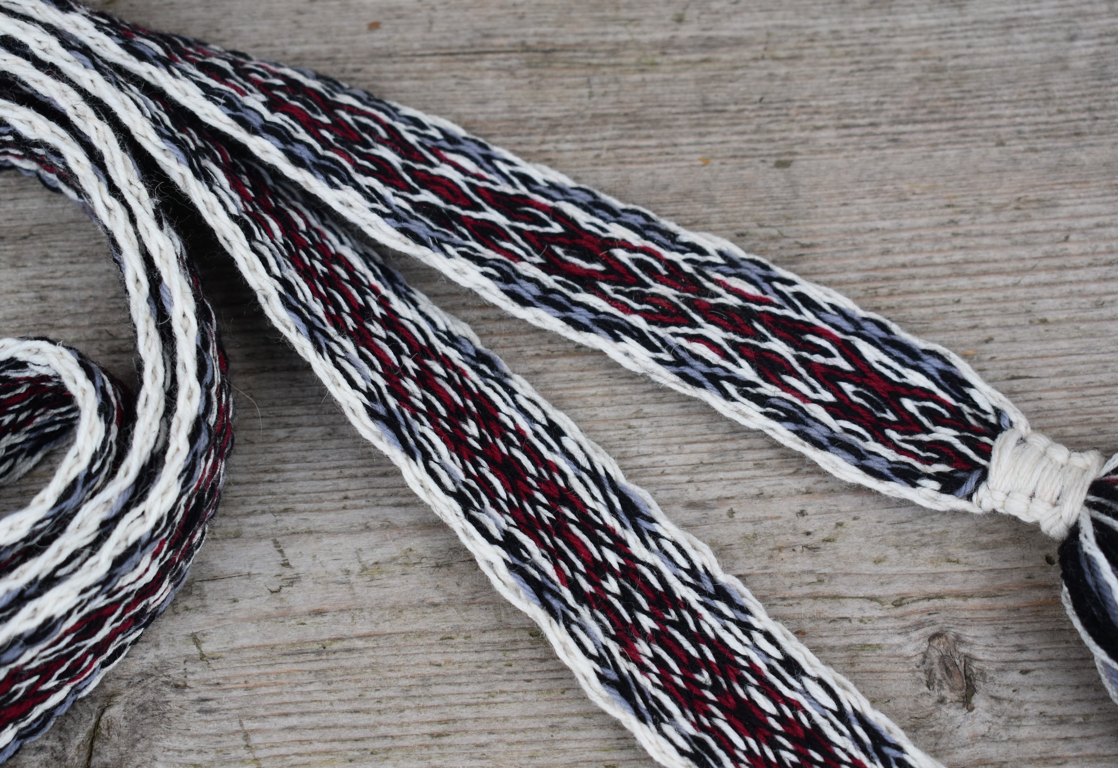 A close-up of a braided textile strap or laces with alternating stripes of white, dark navy, and deep red, laid out on a rustic light gray wooden surface.