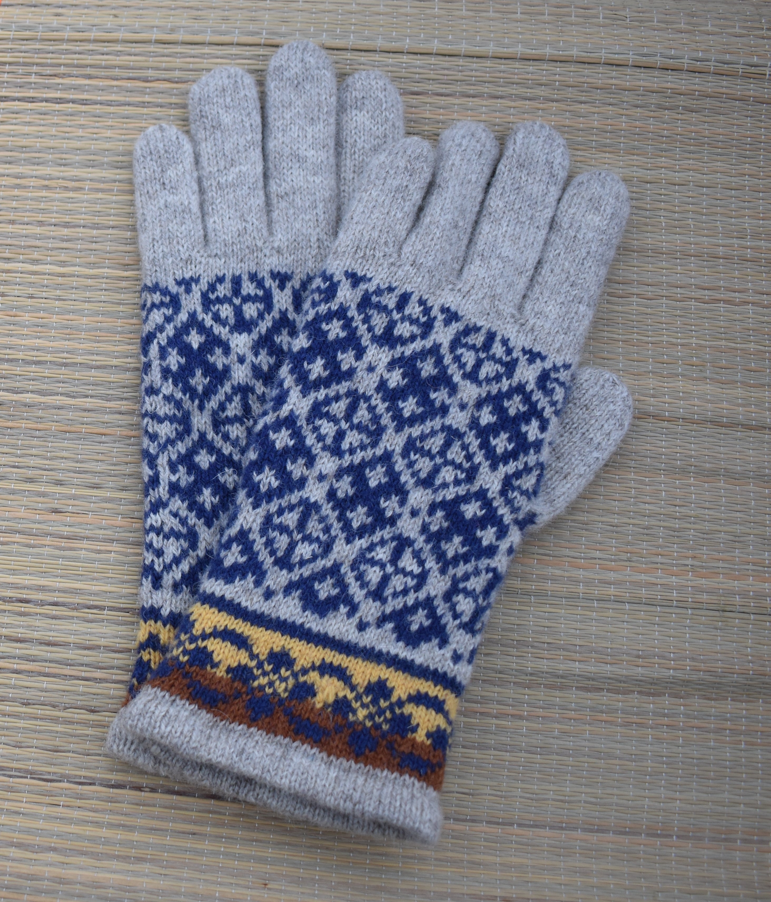 A pair of knit winter gloves in light gray with a blue and white Fair Isle pattern and a yellow and brown patterned cuff, displayed on a light wood background.
