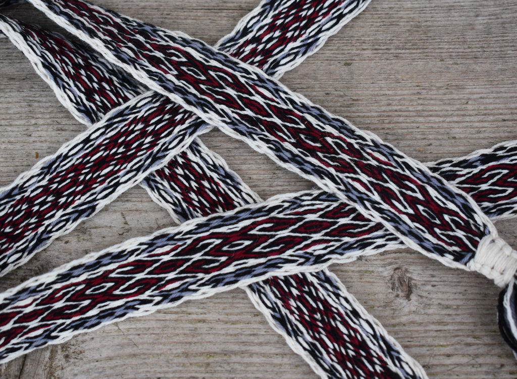 A close-up of braided rope or strap material laid on a weathered wood surface. The product appears woven with white, dark gray, and deep red threads, forming a repeating geometric pattern.