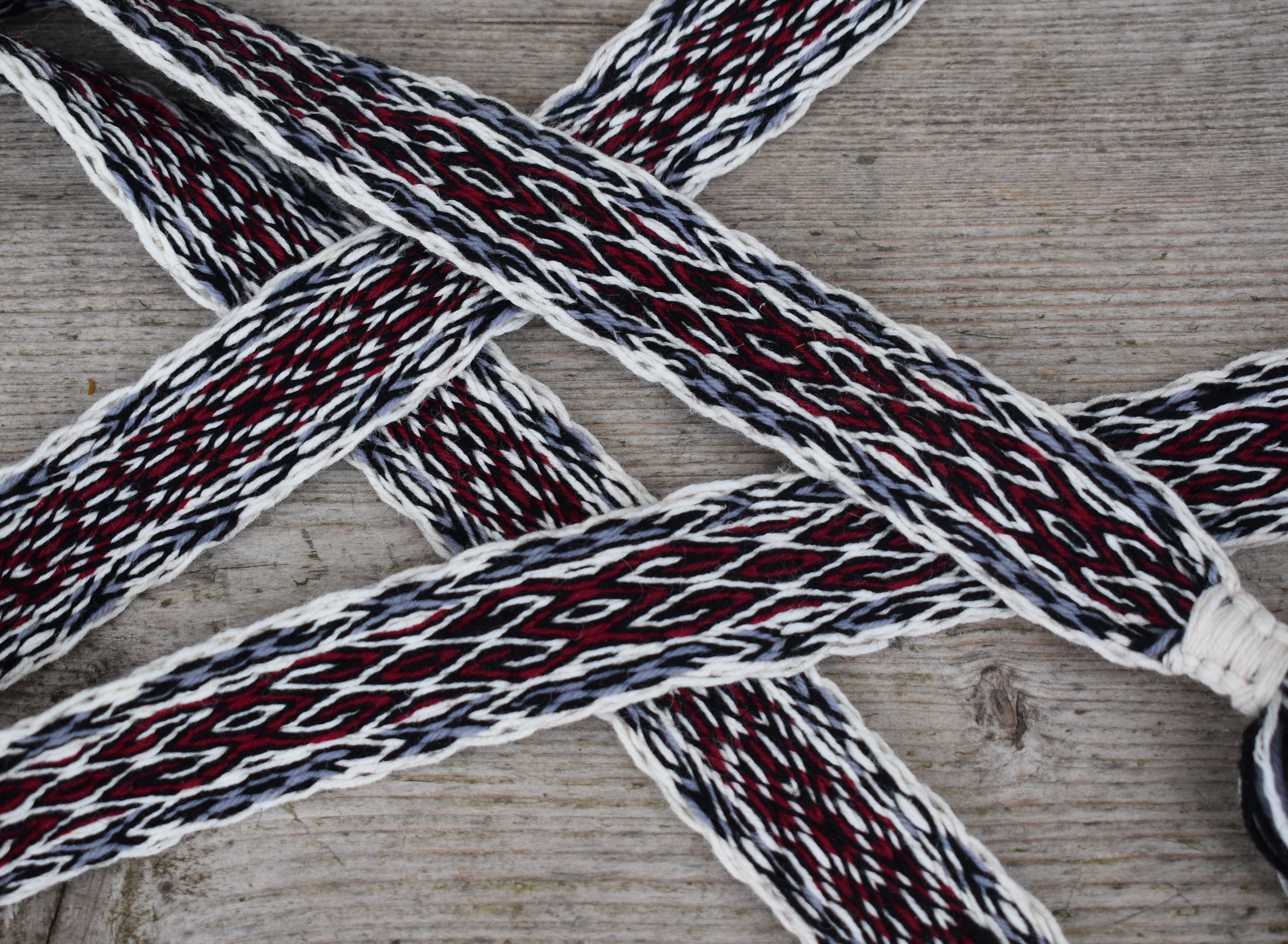 A close-up of braided rope or strap material laid on a weathered wood surface. The product appears woven with white, dark gray, and deep red threads, forming a repeating geometric pattern.