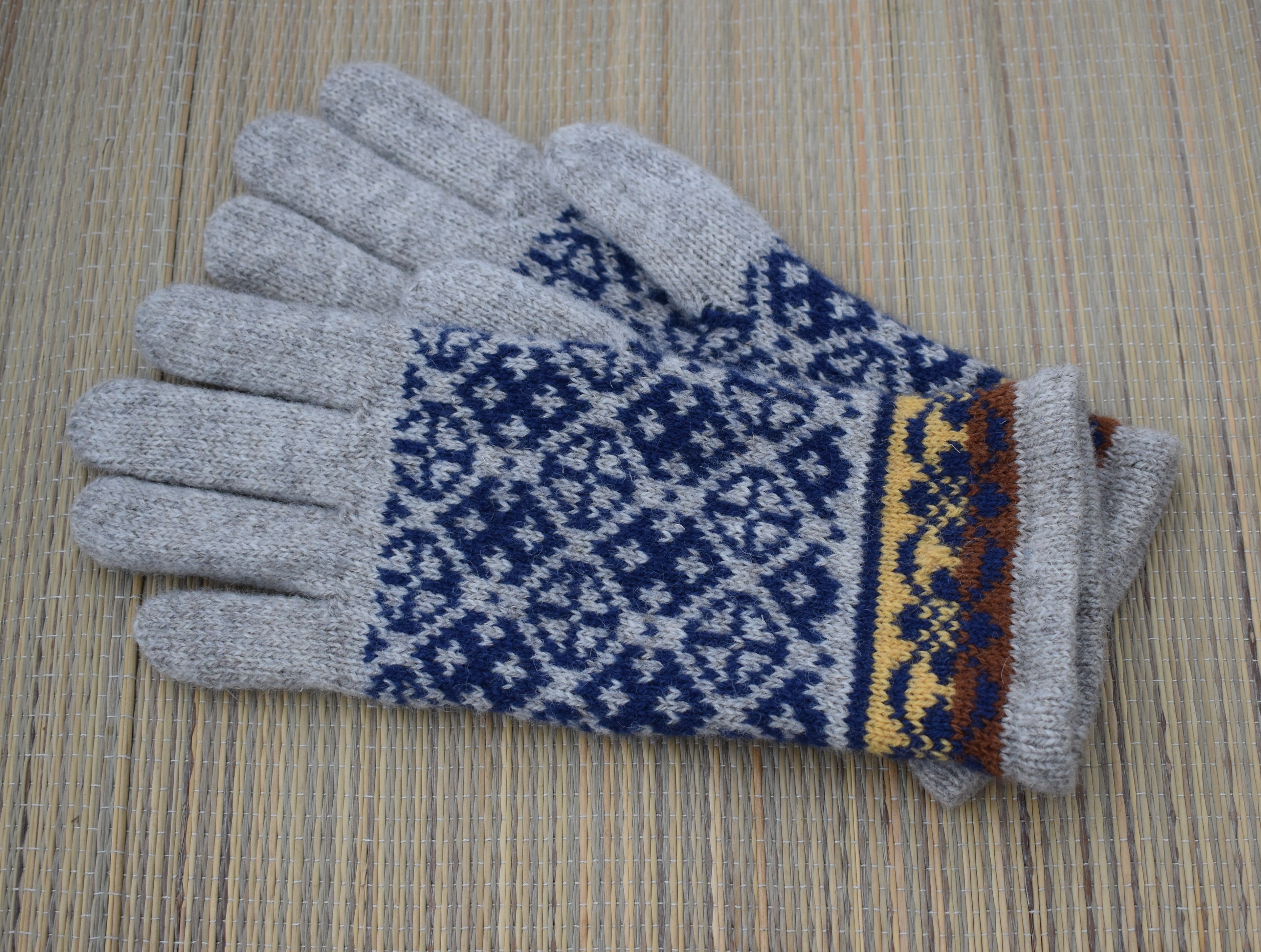 A pair of knit winter gloves with light gray fingertips and backs, featuring a navy blue and white geometric pattern on the palms and a decorative border in yellow and brown near the cuffs, displayed on a woven straw surface.