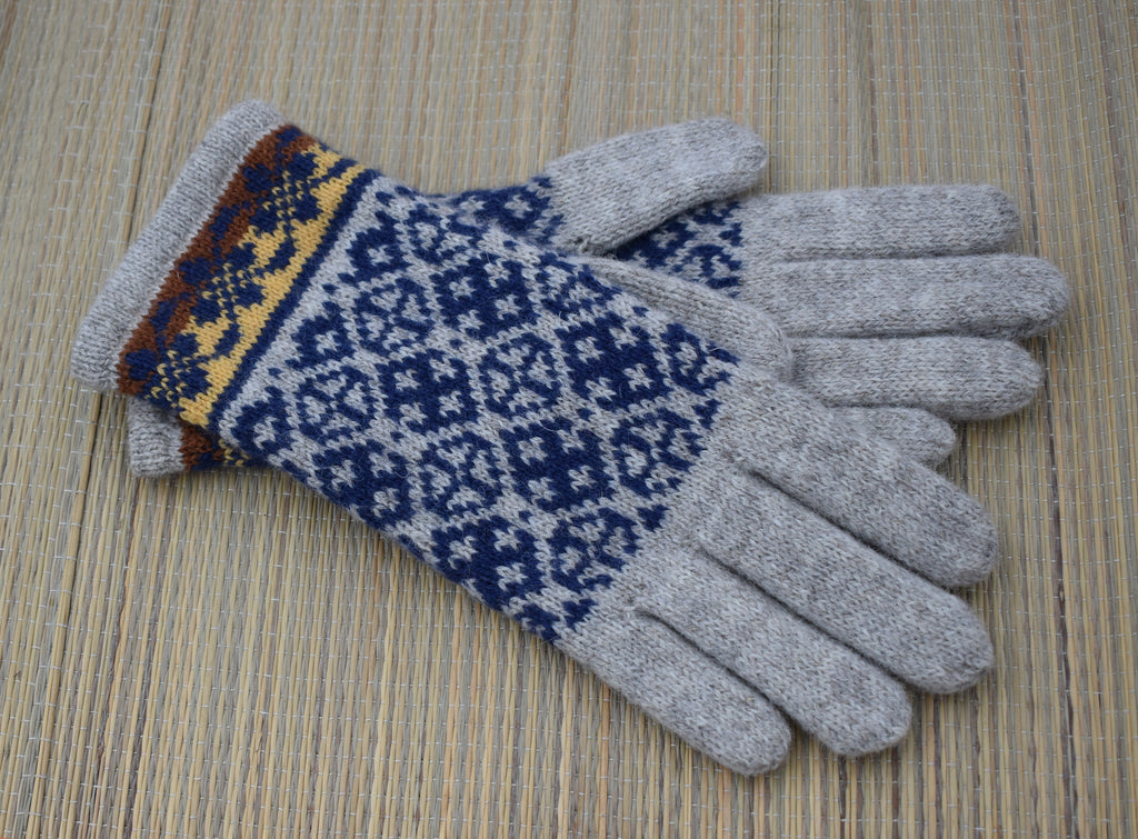 A pair of knit winter gloves with light gray fingertips and palms, featuring a navy blue and white geometric pattern on the backs and a multicolor patterned cuff, displayed on a woven straw mat.