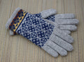 A pair of knit winter gloves with light gray fingertips and palms, featuring a navy blue and white geometric pattern on the backs and a multicolor patterned cuff, displayed on a woven straw mat.