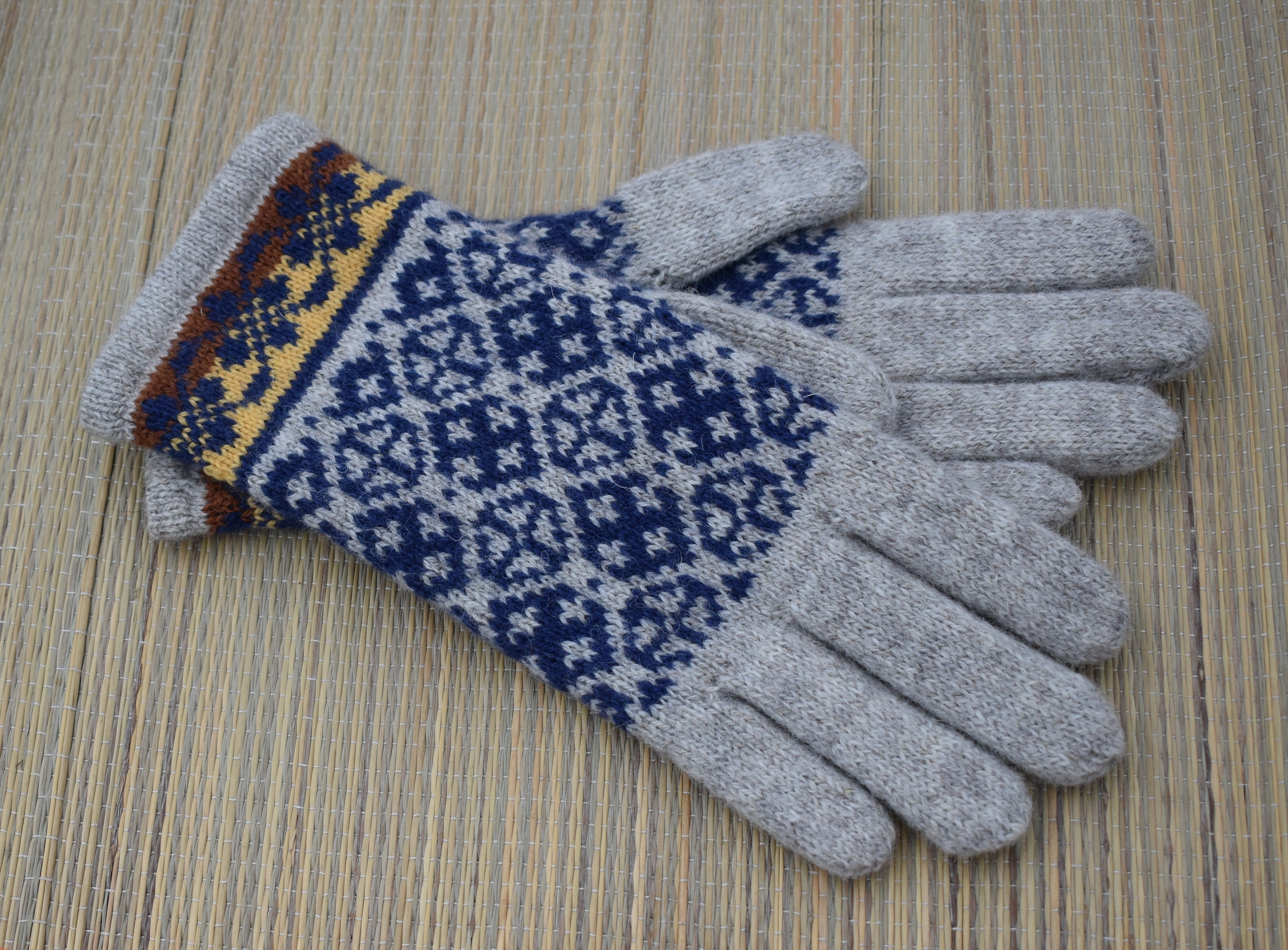 A pair of knit winter gloves with light gray fingertips and palms, featuring a navy blue and white geometric pattern on the backs and a multicolor patterned cuff, displayed on a woven straw mat.