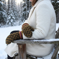 A person sitting in a snowy outdoor chair wearing a cream coat and knit mittens with a yellow and black geometric pattern and red striped trim, with a snowy forest in the background.