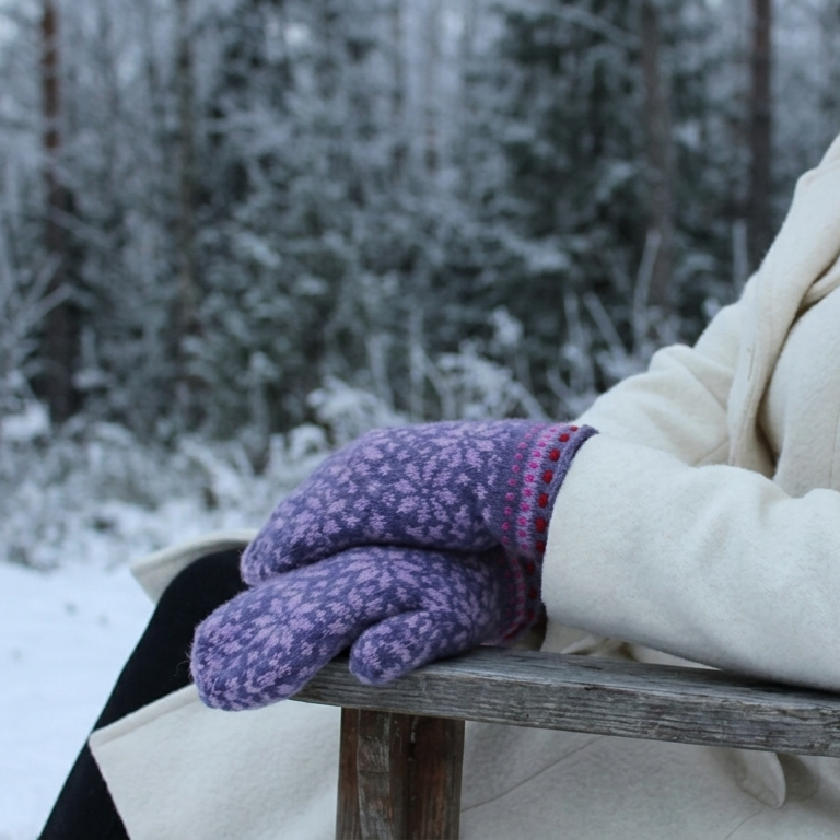 A person seated on a wooden chair in a snowy outdoor setting, wearing a cream coat and purple patterned knit mittens.