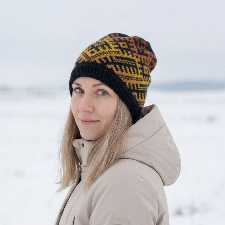 A woman wearing a beige winter jacket and a black, yellow, and orange patterned knit beanie hat, standing in a snowy outdoor landscape.