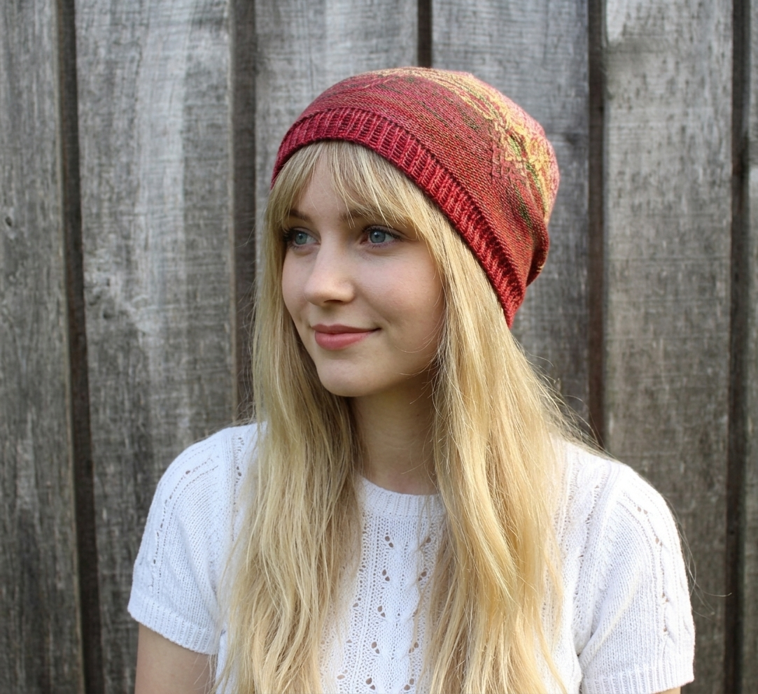 A young blonde woman wearing a red and orange patterned knit beanie hat, standing in front of a weathered gray wooden fence.