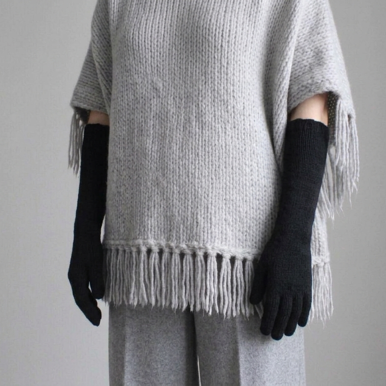 A person wearing a light gray knit poncho sweater with fringe, black knit arm warmers, and gray pants, photographed against a plain light gray wall.