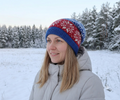 A woman wearing a red and blue knit beanie with a white snowflake pattern, standing outdoors in a snowy landscape with trees in the background.