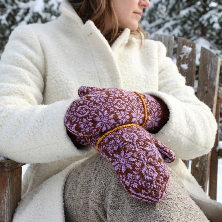 A person sitting outdoors in a snowy setting wearing a cream-colored coat and purple knit winter mittens with a floral pattern and orange trim.