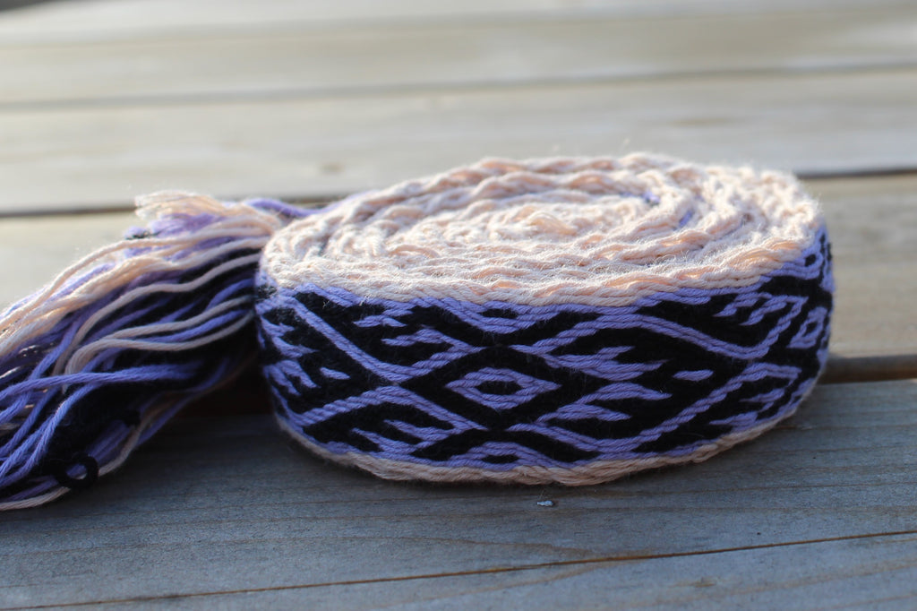 A rolled piece of handwoven cotton braid with a black and lavender geometric pattern and light peach ends, displayed on a weathered gray wooden deck.