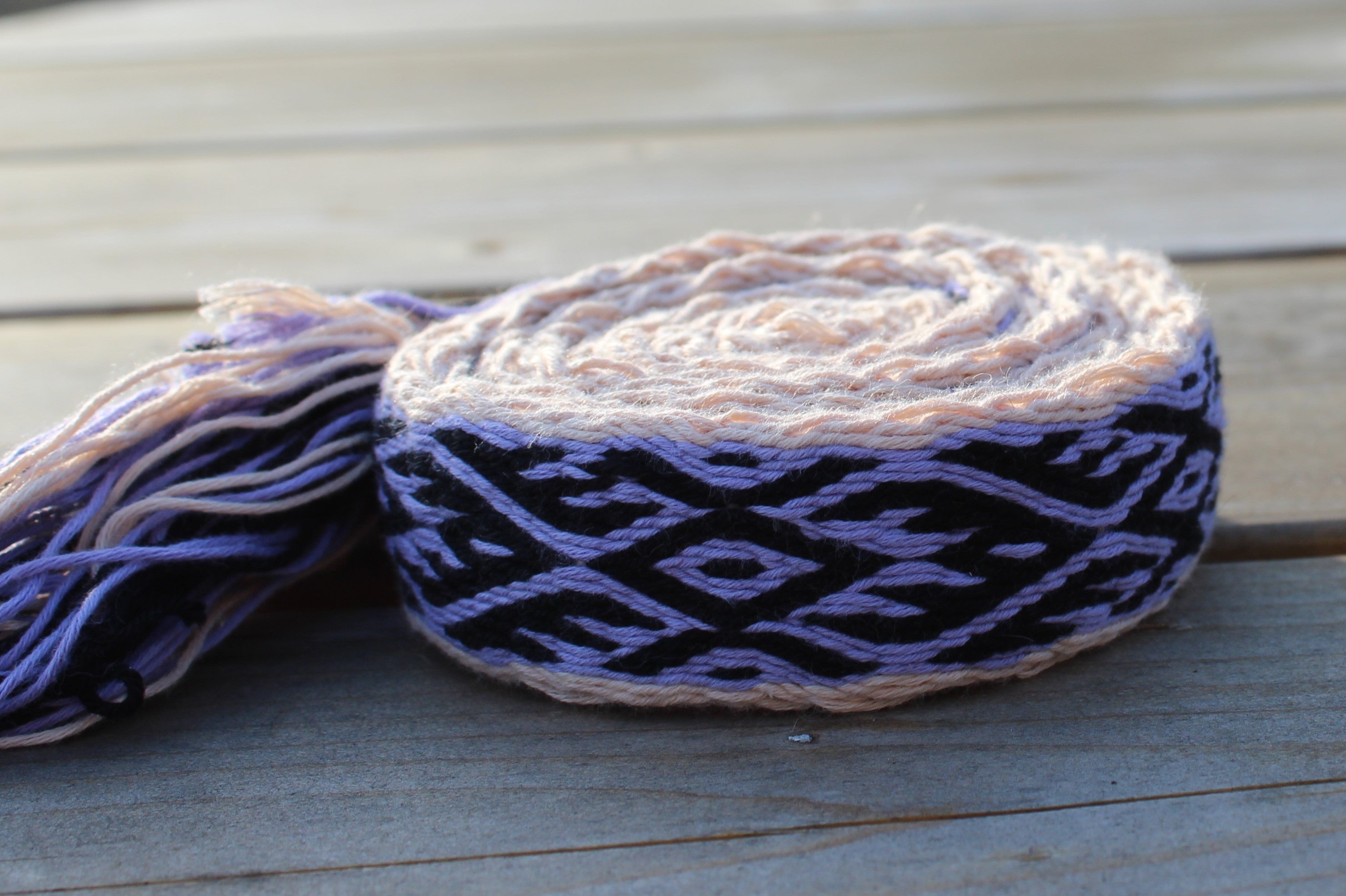 A rolled piece of handwoven cotton braid with a black and lavender geometric pattern and light peach ends, displayed on a weathered gray wooden deck.
