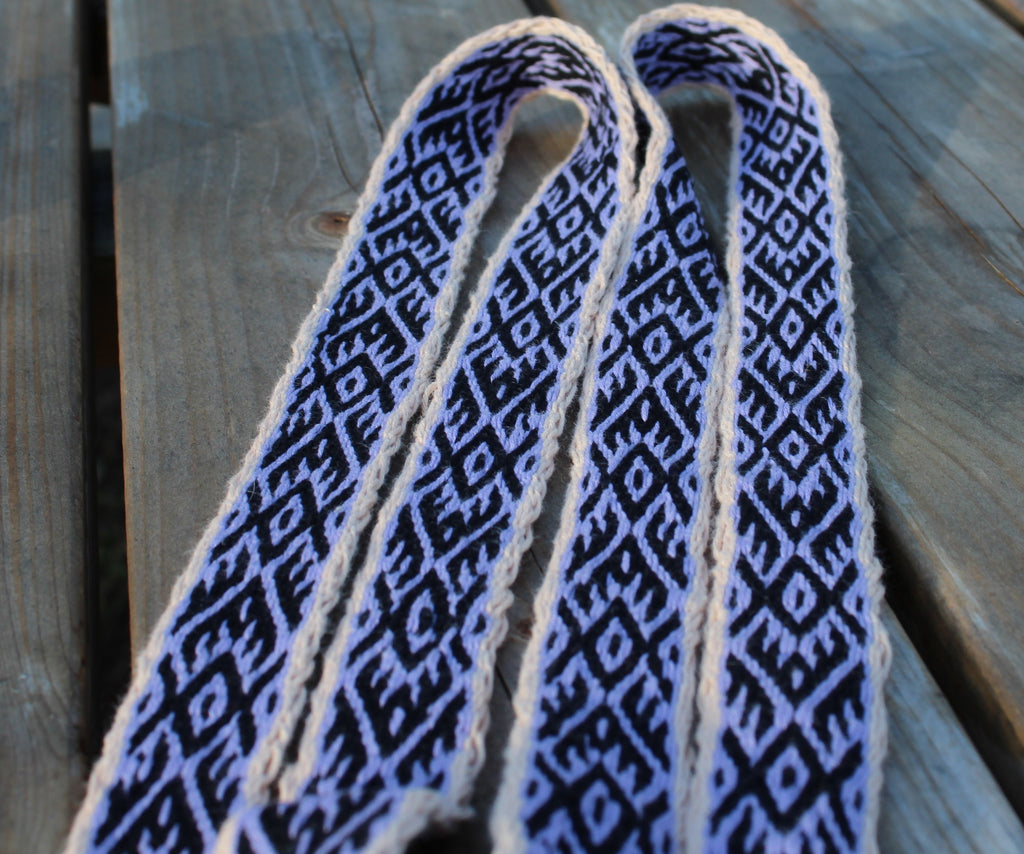 A close-up of a woven strap or belt with a black and light blue geometric pattern and cream edges, laid on a weathered wooden surface.
