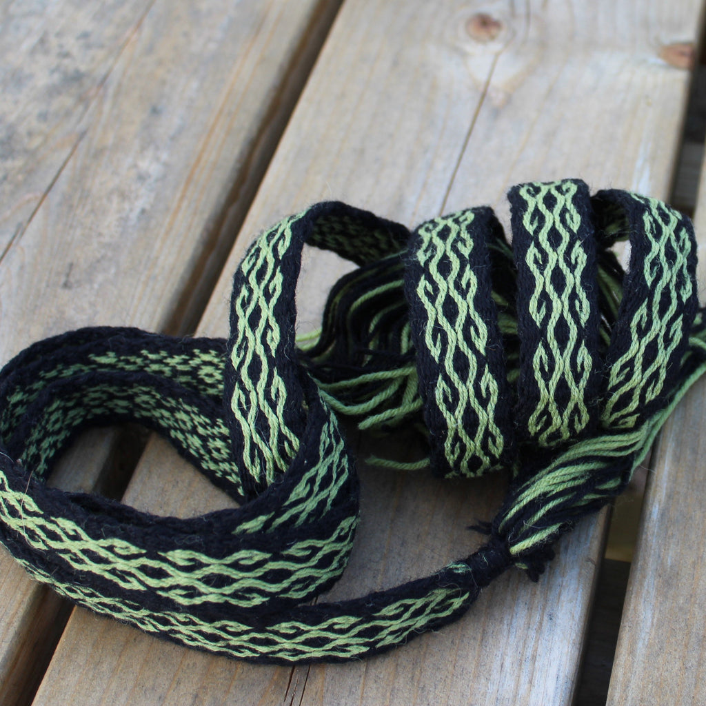 A coiled black and light green woven strap or cord with a braided pattern, resting on a light-colored wooden plank surface.