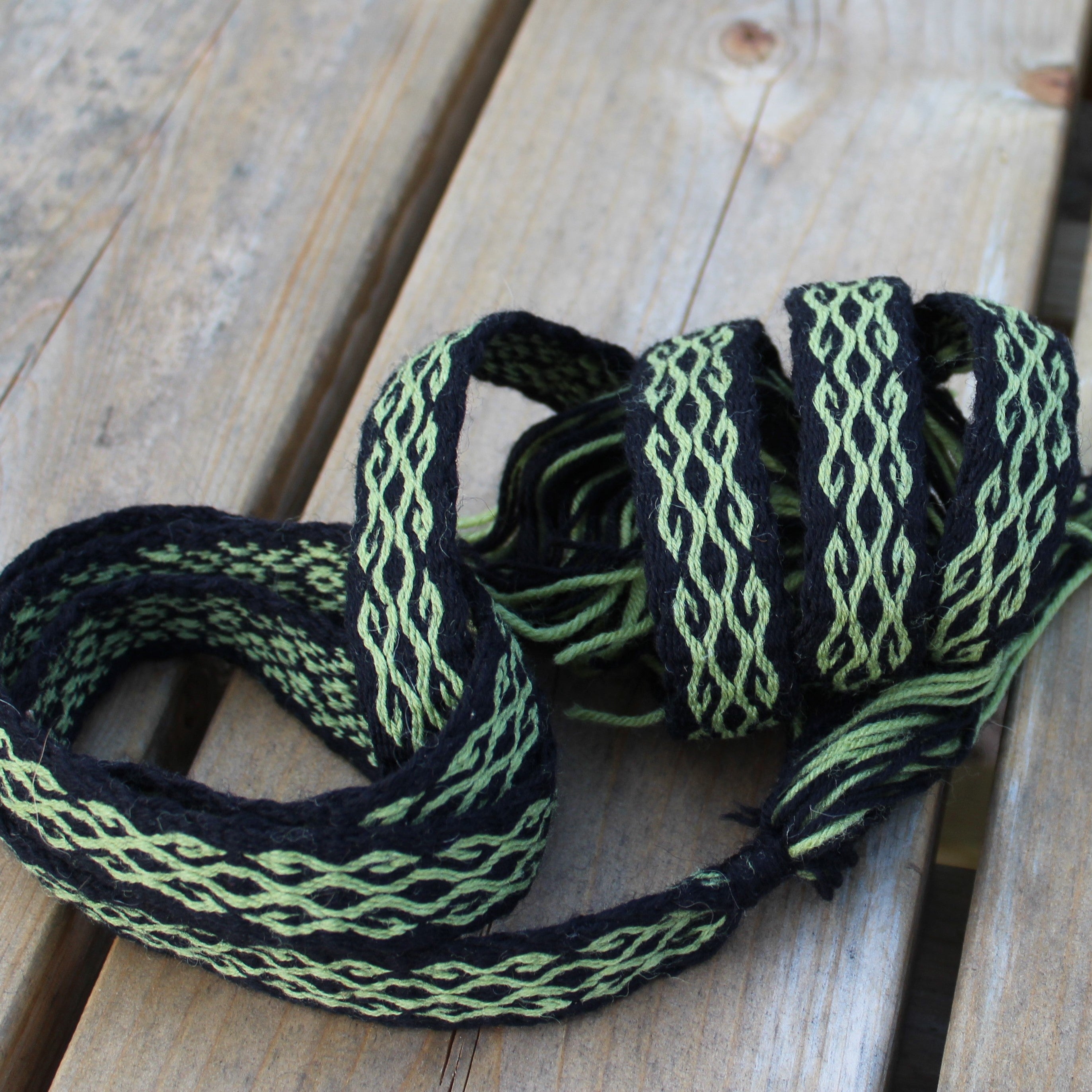 A coiled black and light green woven strap or cord with a braided pattern, resting on a light-colored wooden plank surface.