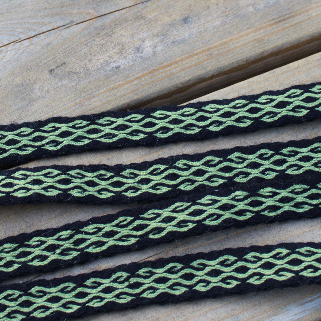 A close-up of a woven black and green braided strap or cord laid diagonally across a light wood surface.