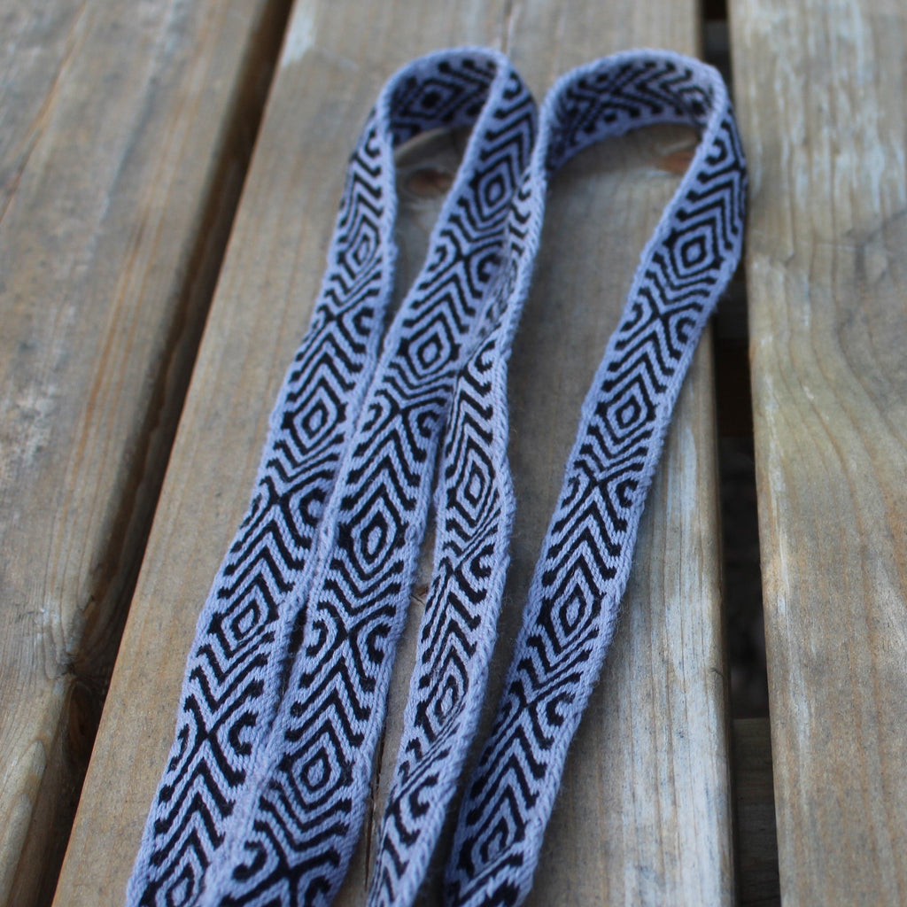 A woven shoulder strap with a black and gray geometric diamond pattern and fringed ends, laid out on a wooden plank surface.