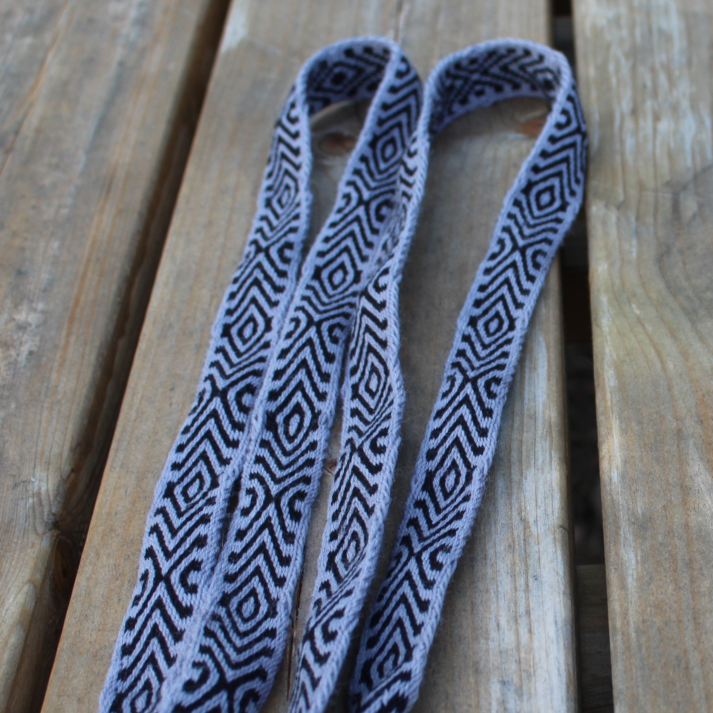 A woven shoulder strap with a black and gray geometric diamond pattern and fringed ends, laid out on a wooden plank surface.
