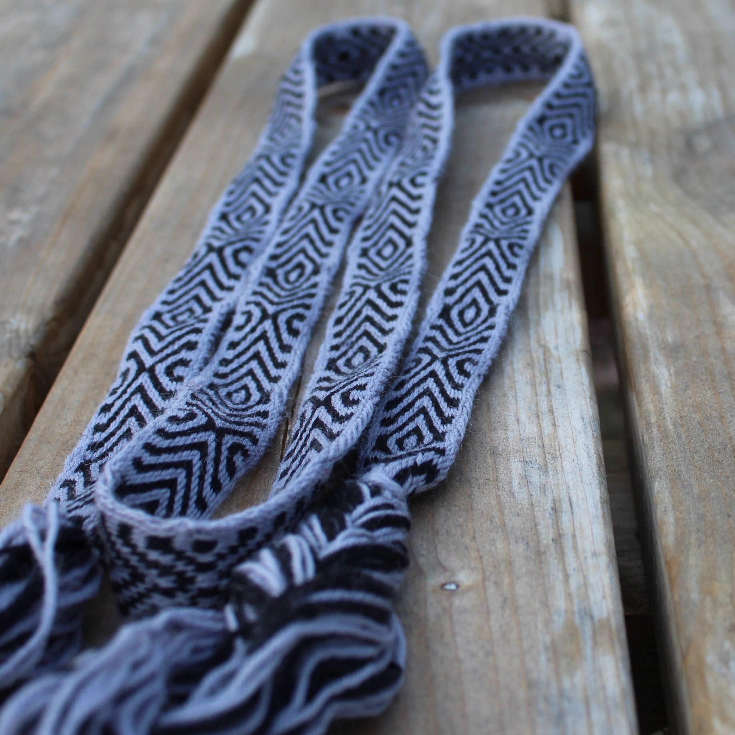 A close-up of a woven textile accessory, likely a strap or belt, featuring a black and gray geometric pattern on a light gray base, resting on weathered wooden planks.