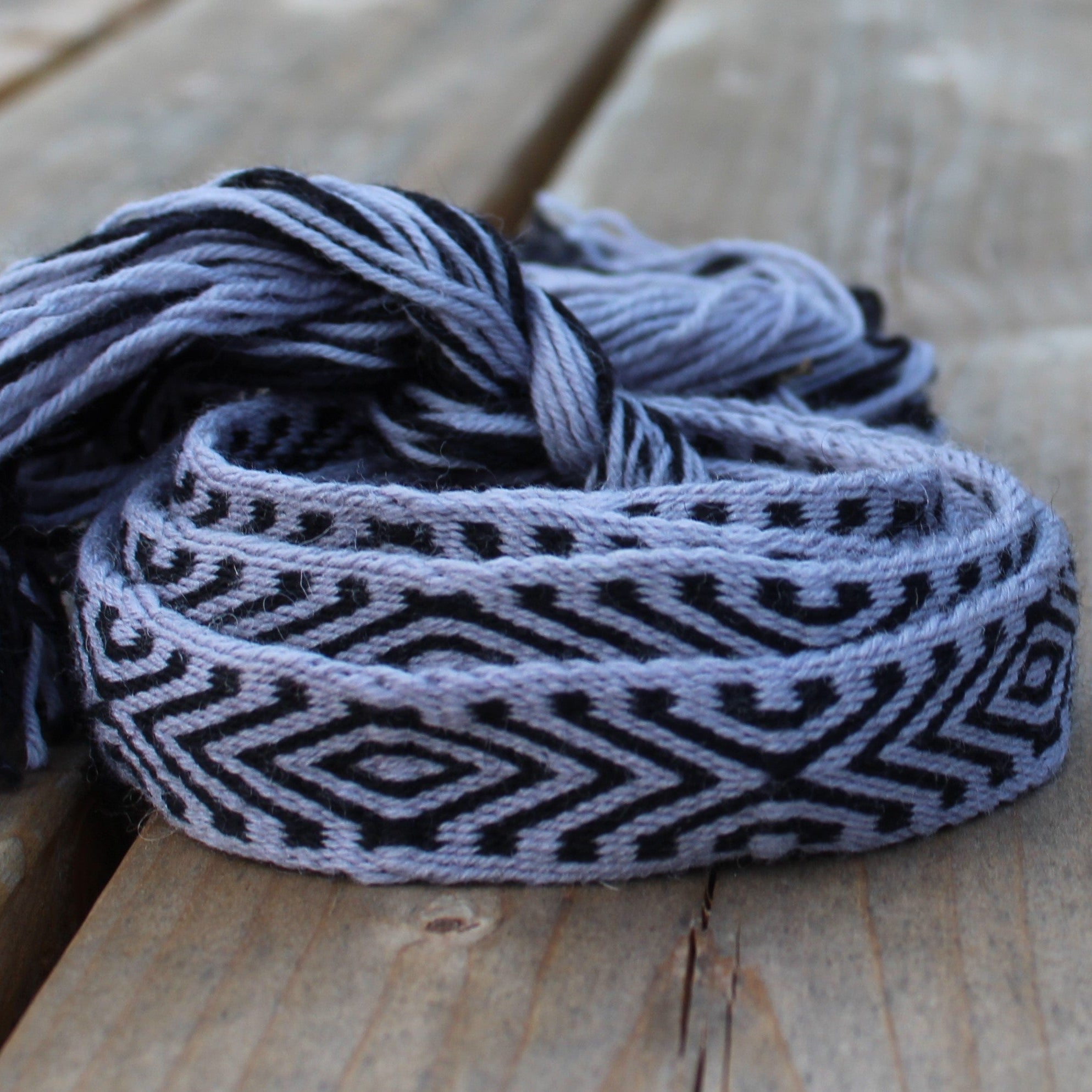 A woven belt with a black and light gray geometric diamond pattern and fringed ends, resting on a rustic wooden plank surface.