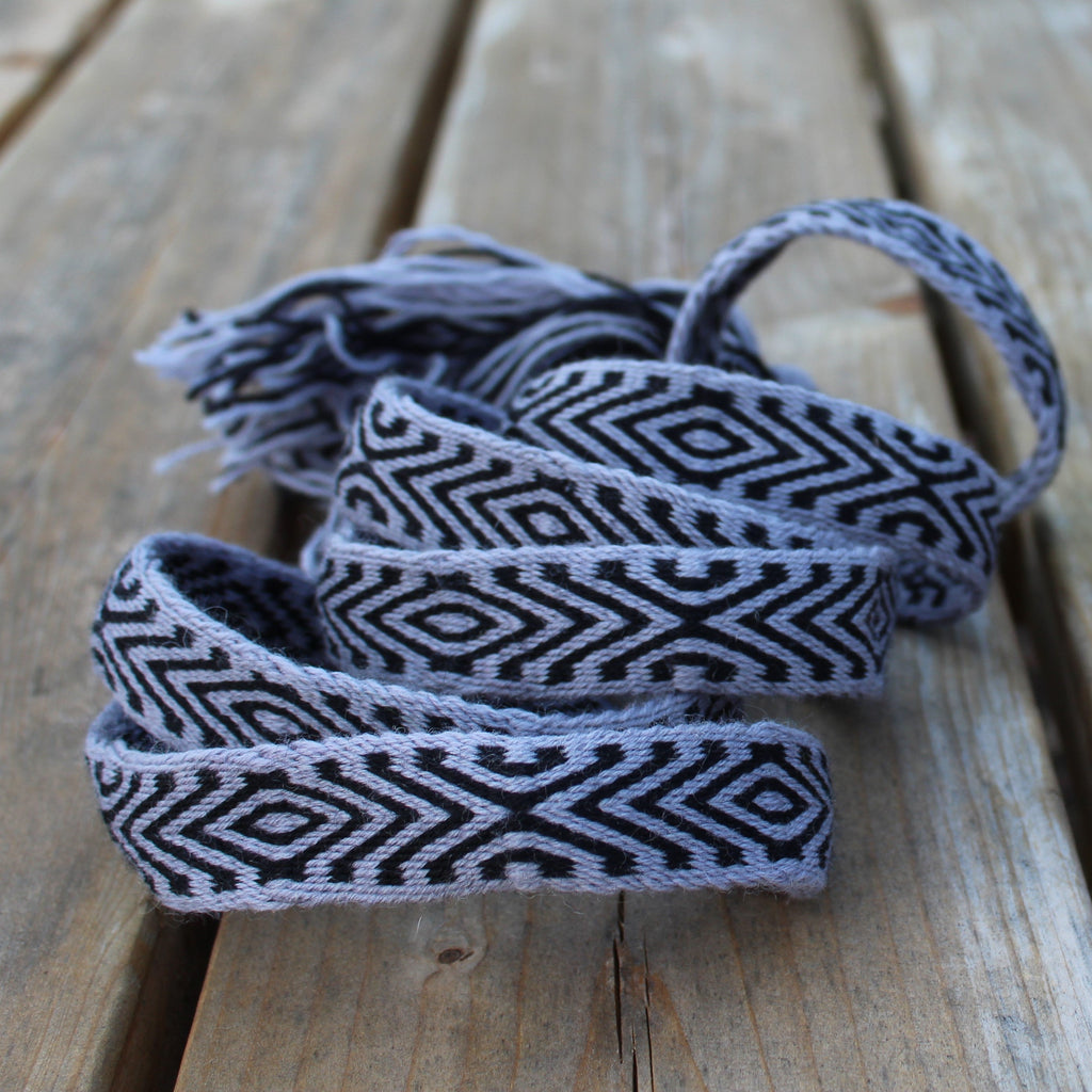 A coiled piece of woven ribbon with a black and light gray geometric diamond pattern, resting on a rustic wooden plank surface.